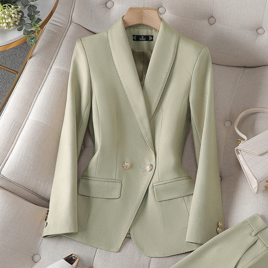 Ein hellgrüner Blazer mit maßgeschneidertem Design und goldenen Knöpfen, präsentiert auf einer weichen Oberfläche neben einer kleinen weißen Handtasche.
