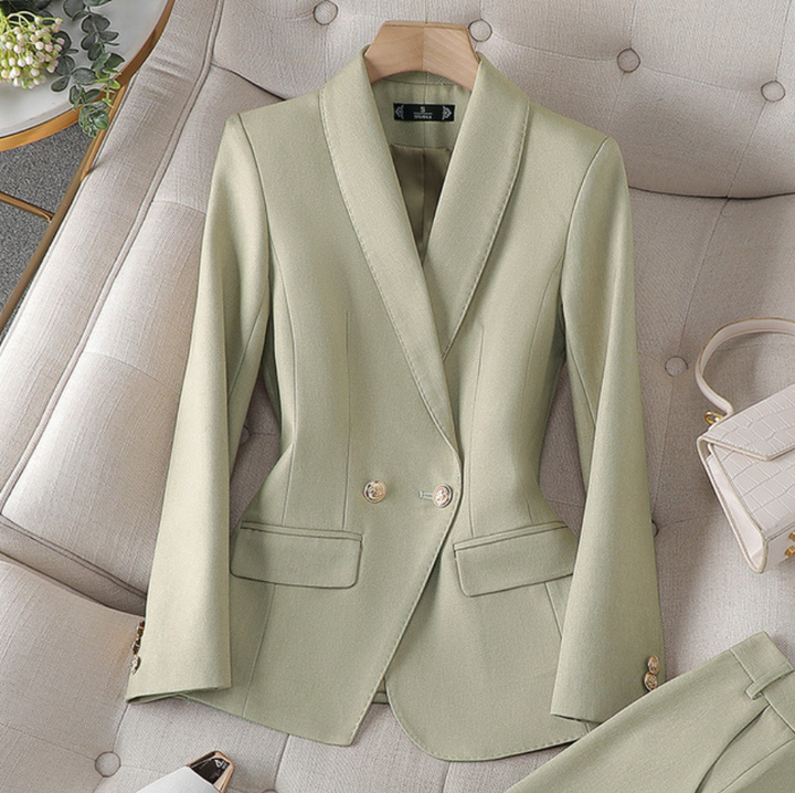 Ein hellgrüner Blazer mit maßgeschneidertem Design und goldenen Knöpfen, präsentiert auf einer weichen Oberfläche neben einer kleinen weißen Handtasche.