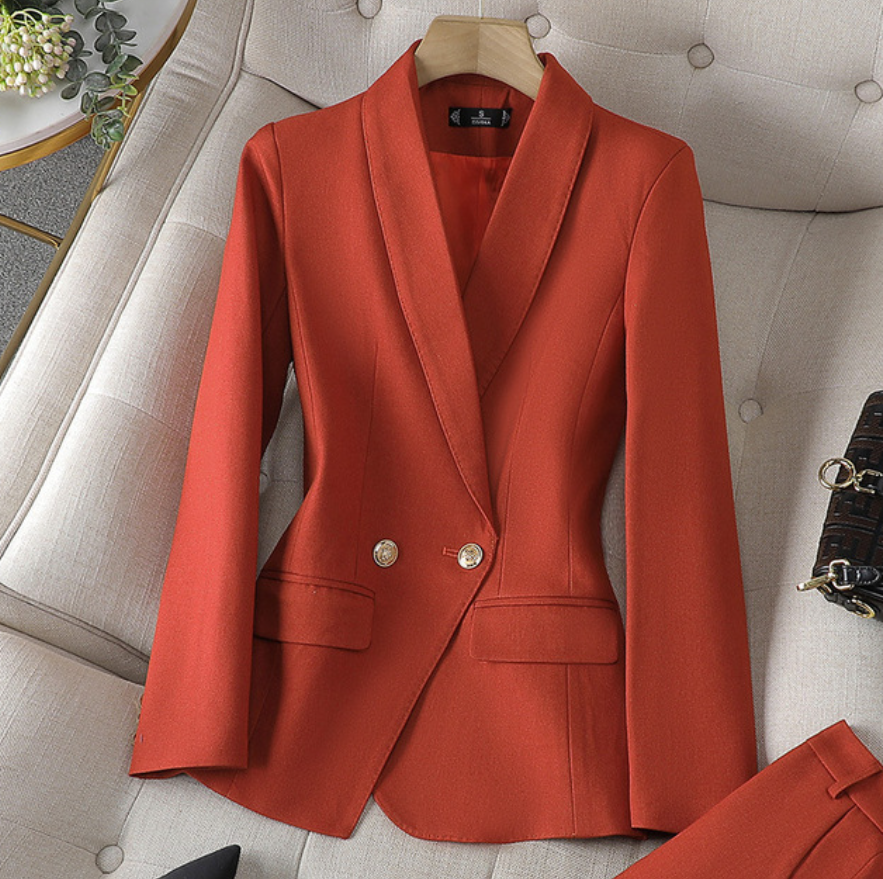 Ein leuchtend gold-orangefarbener Blazer mit maßgeschneiderter Passform und goldenen Knöpfen, präsentiert auf einer weichen Oberfläche neben einer passenden Hose und einer kleinen Handtasche.