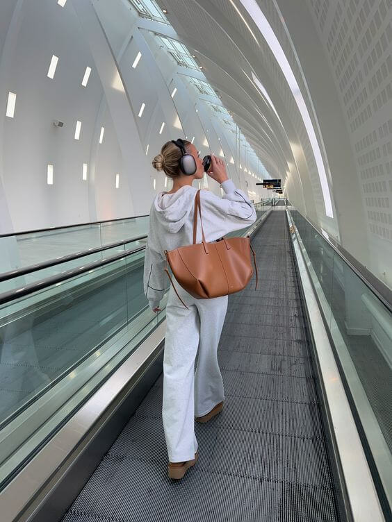 Eine frau in einem grauen outfit und kopfhörern geht durch einen modernen flughafenflur und trägt eine braun ledertasche