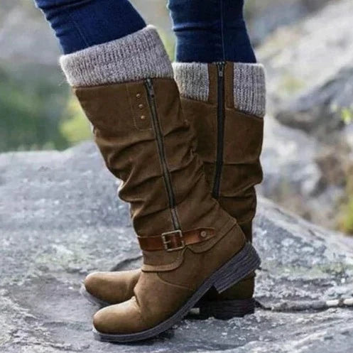 A close-up of a brown orthopedic boot with a casual design and knitted cuff, worn by a person standing on rocky ground, highlighting its stylish appearance and comfort features.